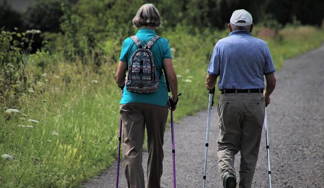 Foto von einem älteren Paar von hinten, das mit Wanderstöcken einen Weg entlang geht.