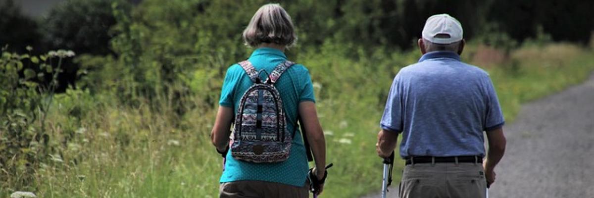 Foto von einem älteren Paar von hinten, das mit Wanderstöcken einen Weg entlang geht.